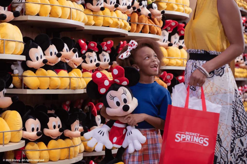 Shopping at Disney Springs A young girl holding a large Minnie Mouse plush looking up at a woman. They are standing in front of a wall of Disney plushes