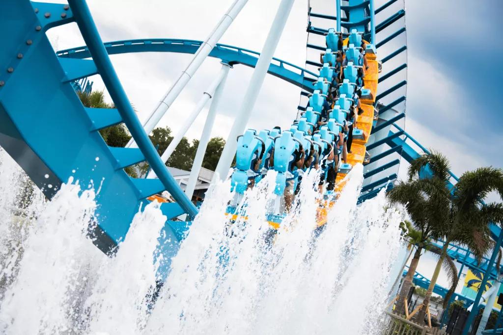 Pipeline: The Surf Coaster A yellow ride vehicle on a blue rollercoaster track heading away from the camera as water rises up from fountains