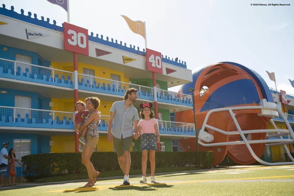 Disney's All-Star Sports Resort A family of four walking past a motel-style hotel building on grass designed to look like an American football field. There is a giant American football helmet in the background.
