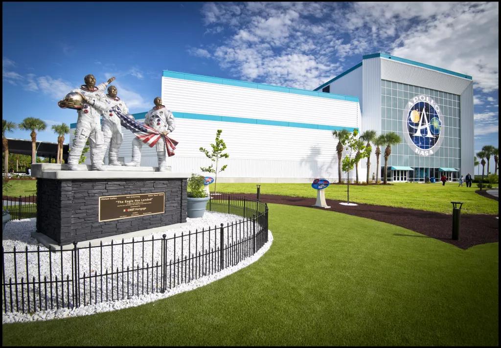 Apollo V Center at Kennedy Space Center A statue of three astronauts holding an American flag in front of a building with a large sign reading Apollo NASA