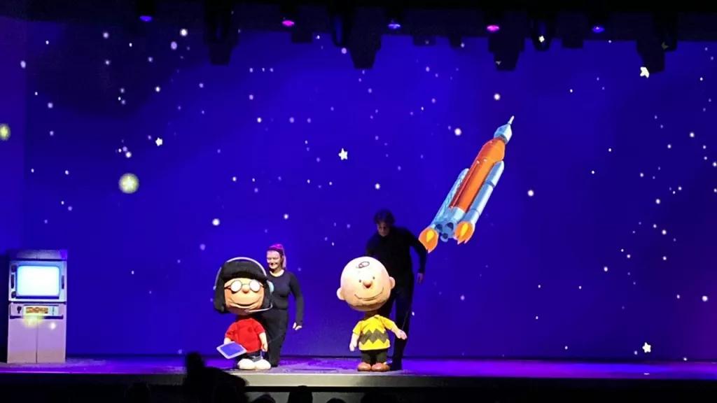 All Systems Are Go at Kennedy Space Center Two people on stage holding life-size puppets depicting Charlie Brown and Lucy from the Peanuts comics
