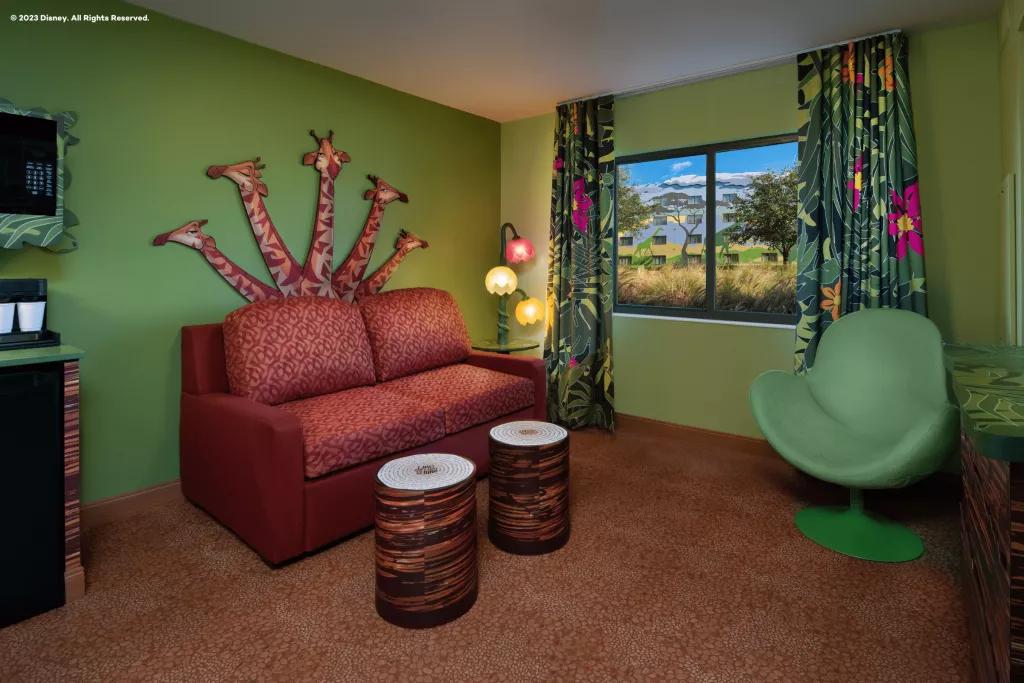 Lion King Suite at Disney's Art of Animation Resort A living area in a hotel suite with a giraffe mural on the wall behind a red sofa and leaf-print curtains on the window