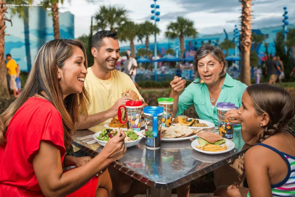 Disney Refillable Mugs A family of 3 adults and 1 child eating sandwiches and salads outside and drinking from reusable mugs