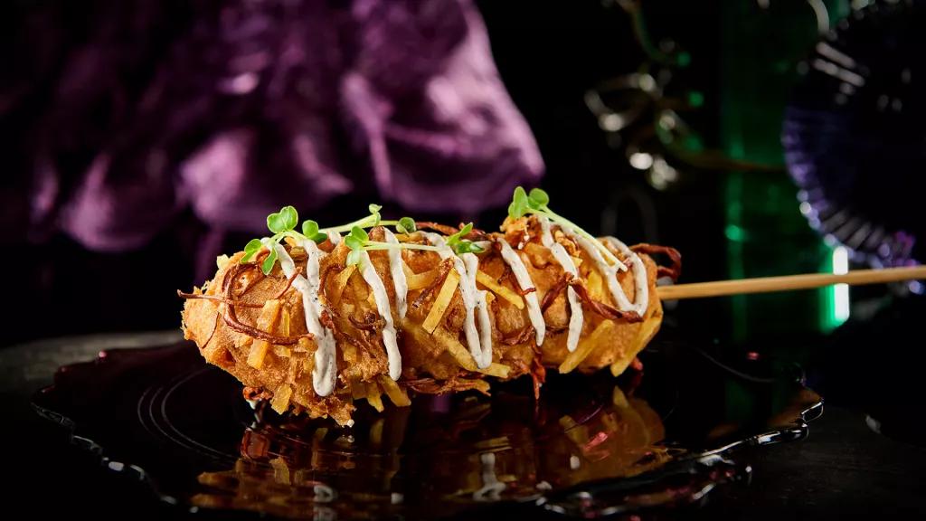 Cordyceps Corndog A corndog on a black plate covered in potato straws, mayo and herbs
