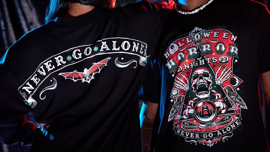 Halloween Horror Nights Apparel A woman facing away from the camera wearing a shirt that reads 'Never Go Alone' and a man facing the camera wearing a tshirt reading 'Halloween Horror Nights'