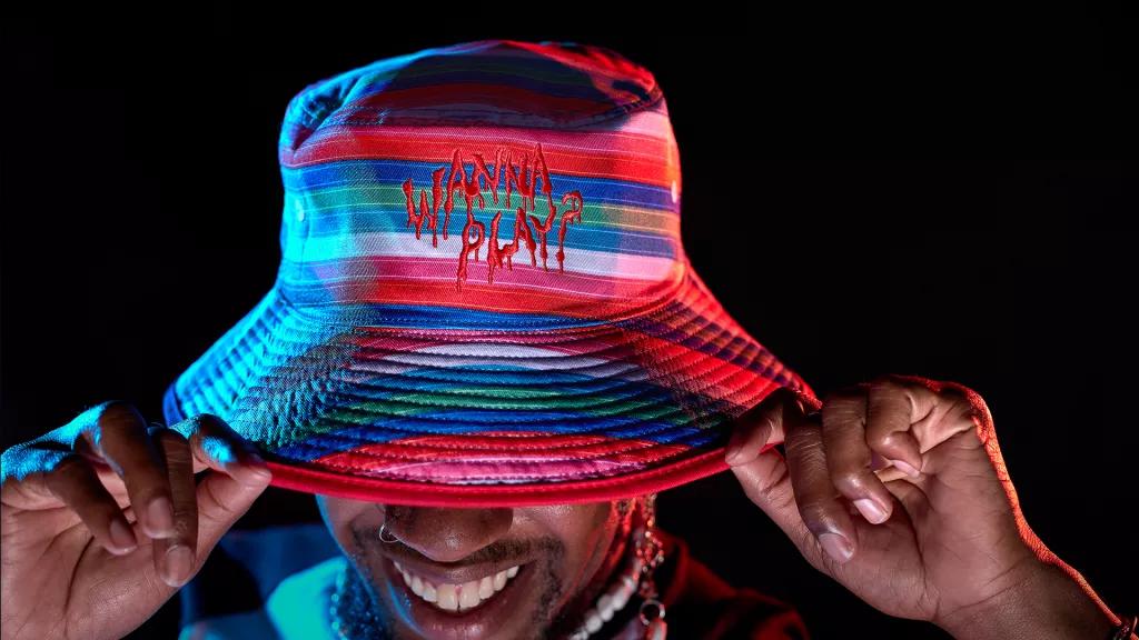 Chucky Apparel A man looking down holding the brim of a striped bucket hat that read's 'Wanna Play?'