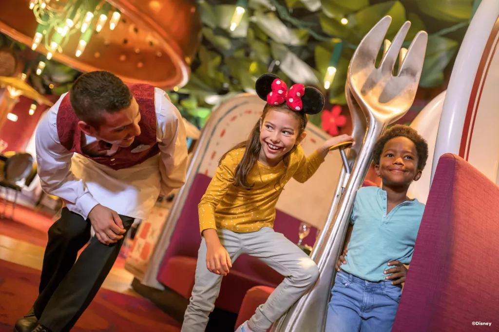 Bistrot Chez Remy A waiter in a red waistcoat smiling at two kids posing next to a larger than life fork