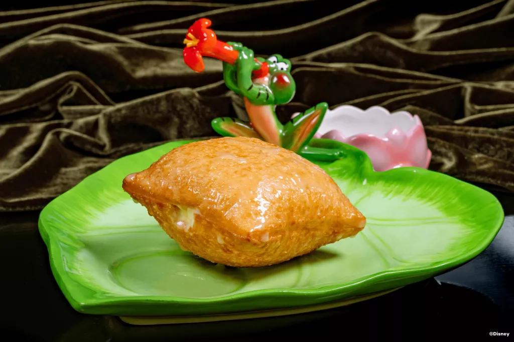 Tiana's Palace House Beignets A filled and glazed beignet on a plate shaped like a lily pad with a frog playing a flower like a trumpet