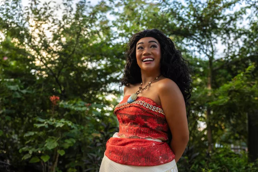 Moana at EPCOT Moana standing with her hands behind her back in front of a group of trees