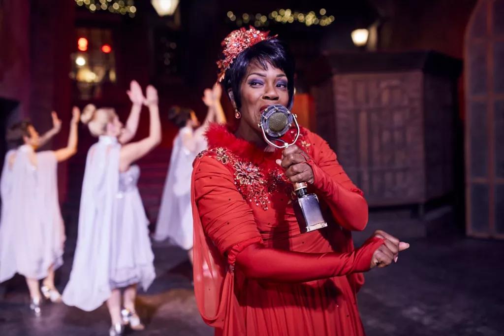 Celestina Warbeck and the Banshees A woman in a red dress singing into an old fashioned microphone. Three women in white dresses are dancing behind her