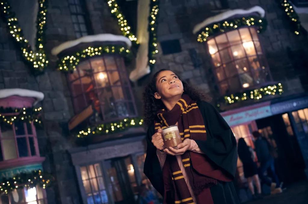 Warm Butterbeer at the Wizarding World of Harry Potter A woman wearing a black Hogwarts cloak and Gryffindor scarf looking up while holding a mug of warm Butterbeer