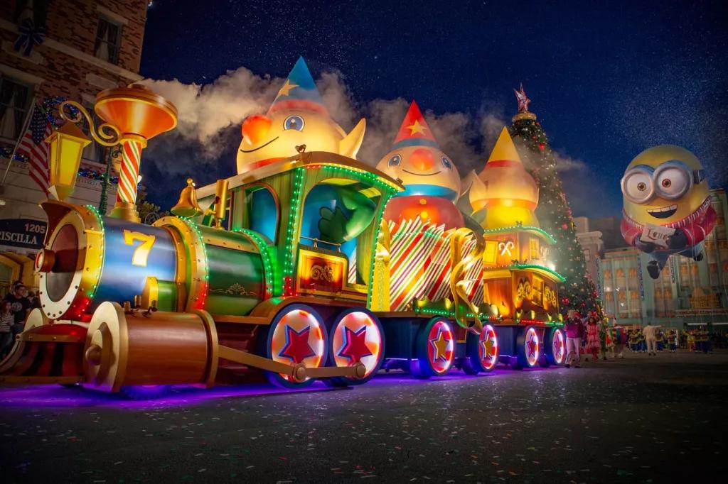 Universal's Holiday Parade featuring Macy's A parade float shaped like a giant toy train lit up with lights. A large balloon shaped like a Minion follows
