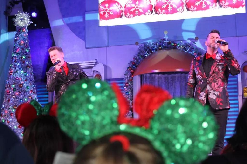 Entertainment at Mickey's Very Merry Christmas Party Two men singing on a stage wearing red and black suits. A pair of sparkly green and red Minnie ears is in the foreground
