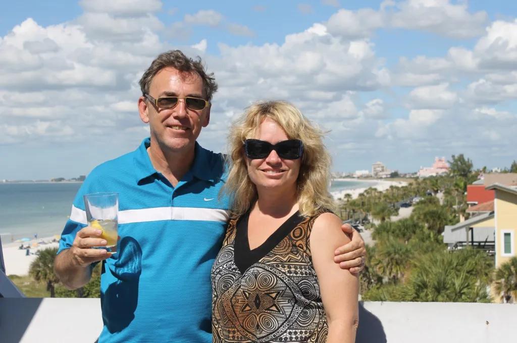 Our Florida Experts - Susan and Simon A man and woman posing on a balcony in front of a shoreline lined with palm trees