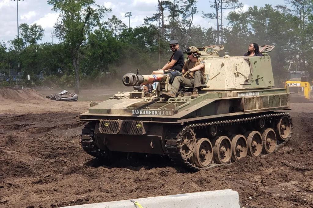 Tank America An army tank being driven across soil with two people sitting at the front and one person at the back