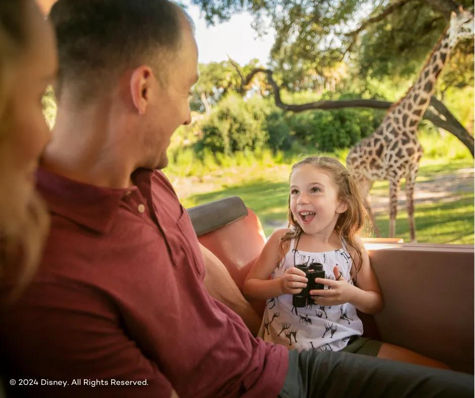 Kilimanjaro Safari at Disney's Animal Kingdom Theme Park Small girl holding binoculars in truck facing her parents with giraffe stood behind her eating leaves in a tree and her dad sat to the left of her smiling down at her.