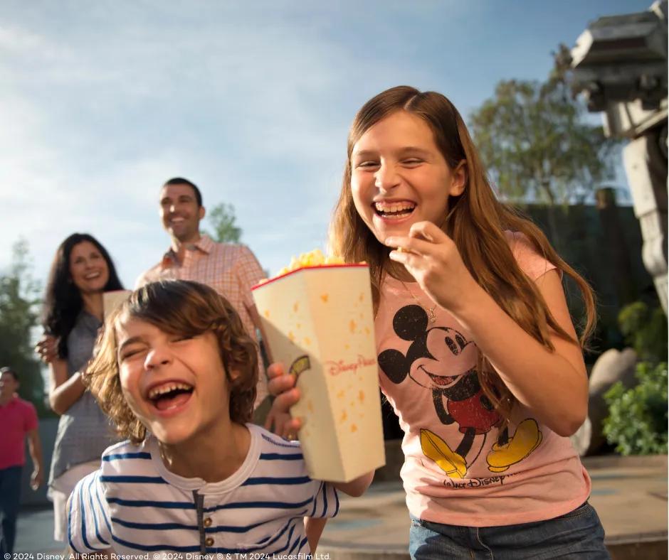 Kids eating popcorn in Disney's Hollywood Studios Girl wearing pink Mickey Mouse top holding popcorn bucket in one hand and a piece of popcorn in the other, laughing with a young boy who is next to her. In the background is a couple smiling and a large star wars robot.