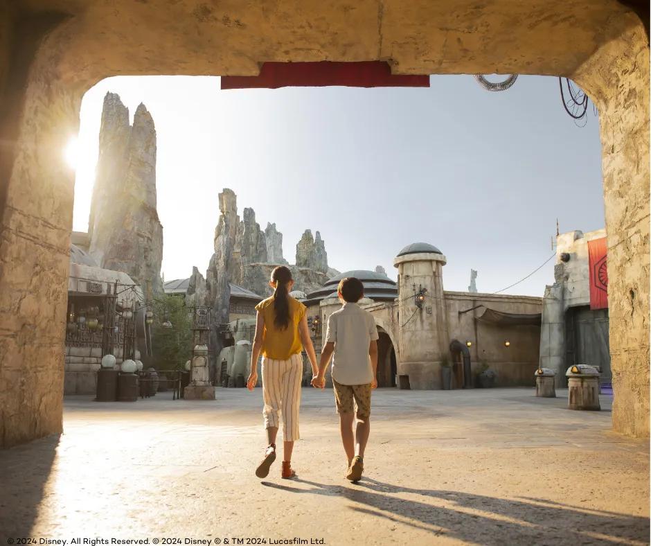 Star Wars: Galaxy's Edge in Disney's Hollywood Studios Young girl and young boy holding hands walking with backs towards the camera, into area themed with grey large rocks and beige dirty buildings with circular roofs. There are wires hanging between the buildings.