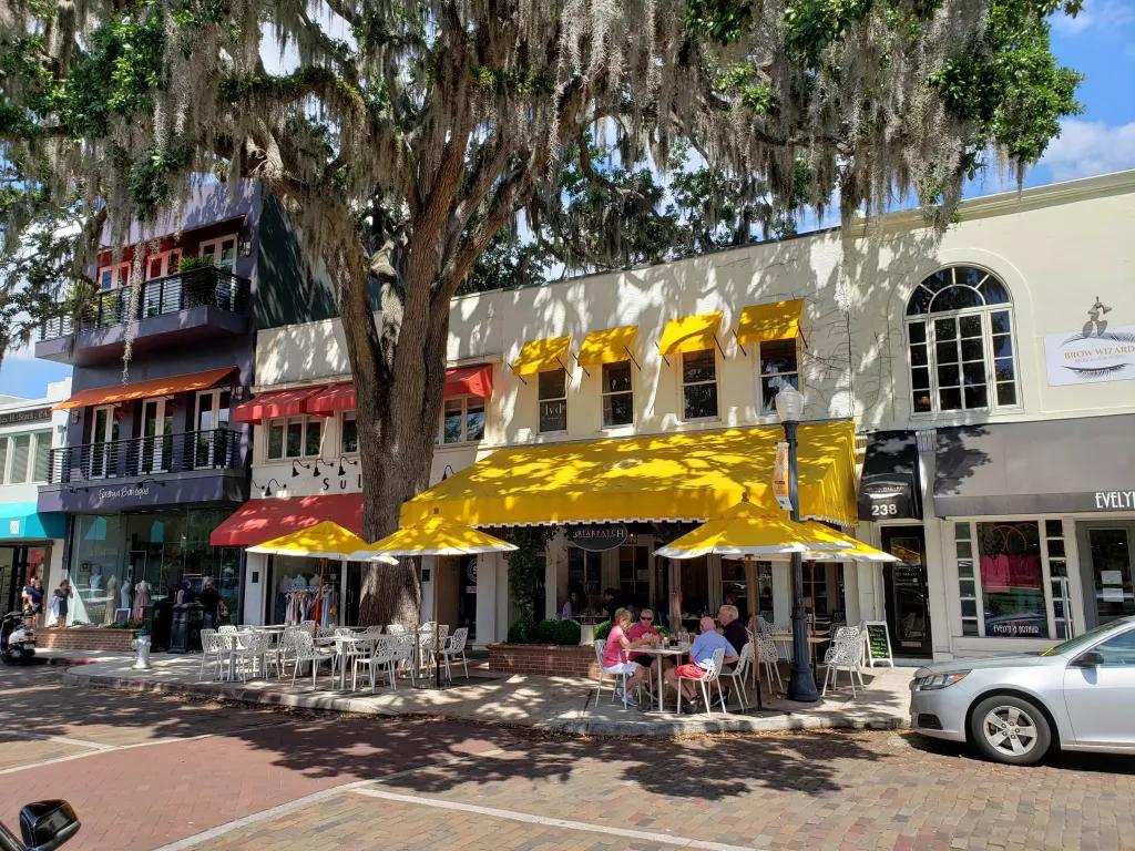 Briarpatch in Winter Park Row of cream coloured buildings with multicoloured shops and restaurants alongside. The central restaurant (Briarpatch) has bright yellow umbrellas outside with white chairs and tables.