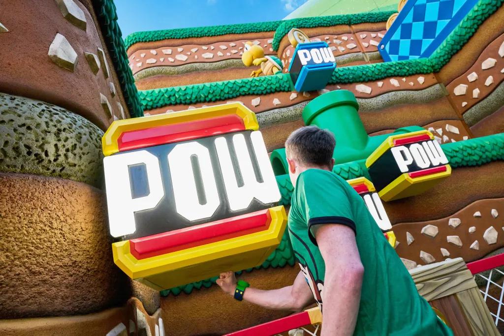 Un homme vêtu d’une chemise verte et d’un groupe de power up vert frappe sous une grande boîte qui flotte et qui dit « POW » en écriture blanche. En arrière-plan se trouve une colline brune avec des décorations de style herbe verte, des tuyaux verts et de petits personnages en forme de tortue.