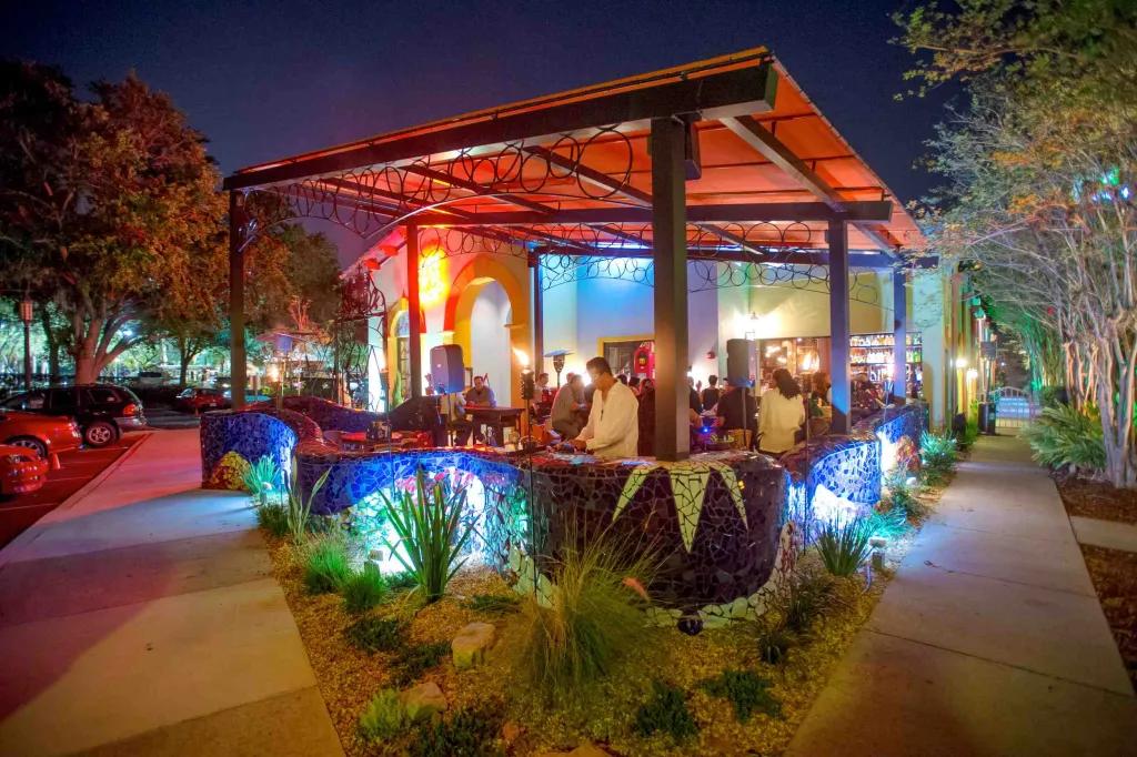 Café Tu Tu Tango - International Drive Large wooden beams holding up an orange roof and a mosaic blue wall winding around the terrace. There are speakers, heaters and people stood on the terrace and there is blue and orange lighting.