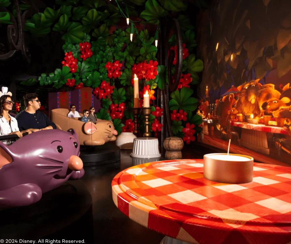 Remy's Ratatouille Adventure in EPCOT Guests sat in a rat shaped vehicle with 3D glasses on. Next to them on the right is a giant bush with large flowers, a giant table with a red and white table cloth, giant candles and a screen with the ratatouille characters on.