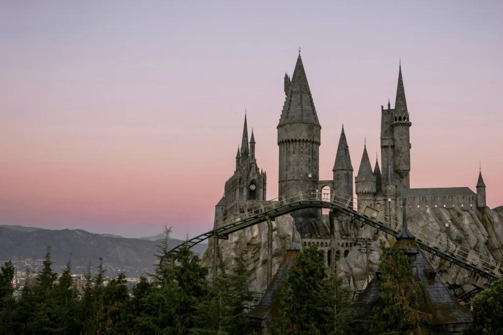 Le château gris de Poudlard était assis sur des rochers avec des arbres et de petits sous-verres devant. En arrière-plan, il y a un coucher de soleil rose et de grandes montagnes.