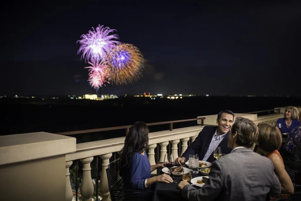 Capa in the Four Seasons hotel Terrace with group of 4 sat around circular table, with a man smiling looking at the others as they watch fireworks being launched in the night sky in the far distance.