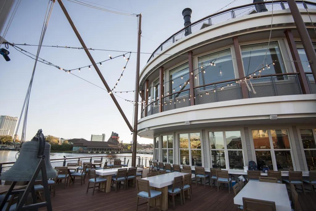 Paddlefish in Disney Springs White tables with 6 wooden chairs around them on a terrace with lake in the background and lights hanging overhead. Also a large bell on the terrace and circular building to the right.