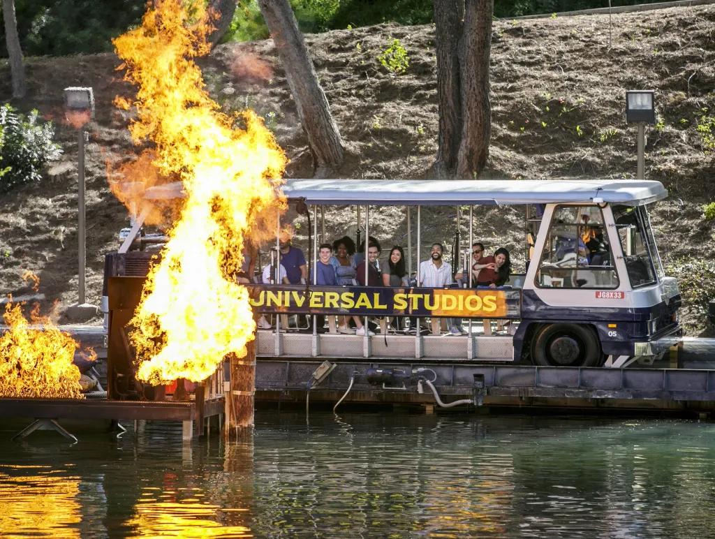Lac avec une grande voiture avec des rangées de personnes, sur le côté de ce véhicule, il est écrit 'Universal Studios'. Au-dessus de l’eau, il y a une jetée en bois qui explose en feu.