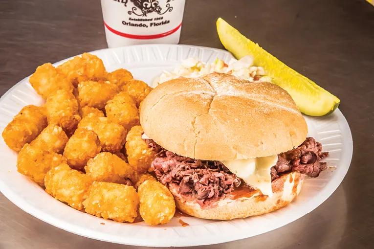 Beefy King Paper white plate with tater tots on and a burger bun with pulled pork/beef inside with a white sauce. In the background there is a white cup.