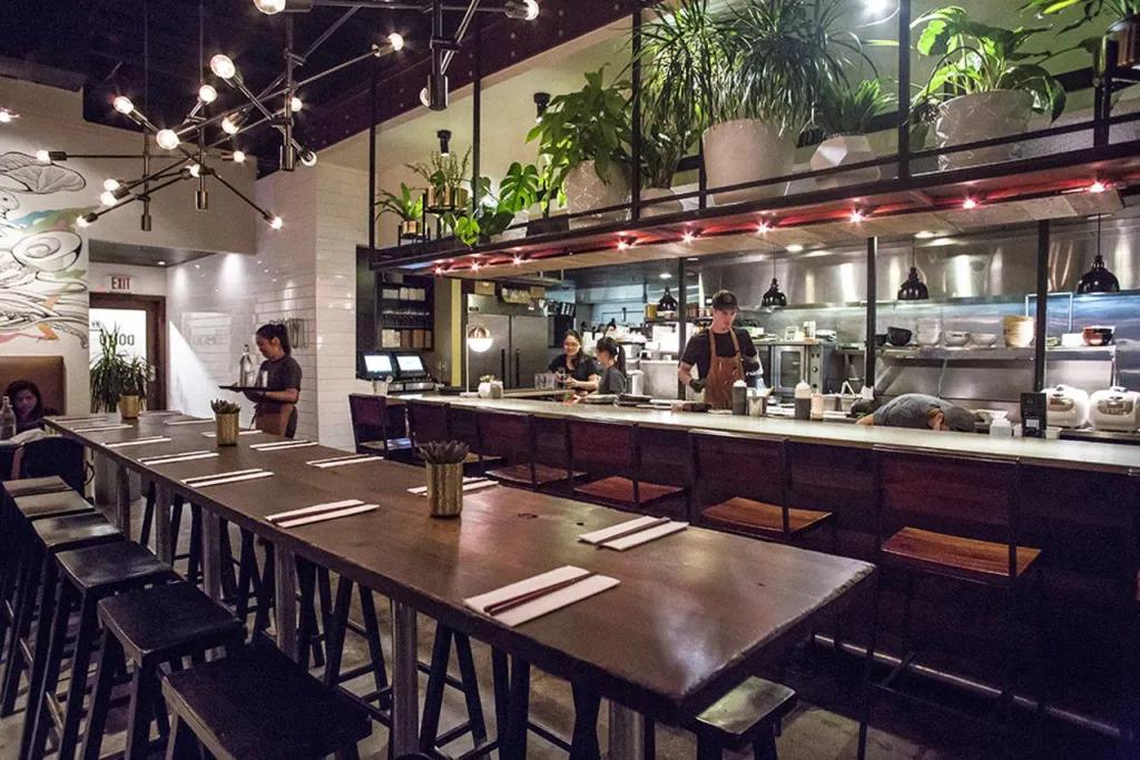 Domu Long wooden table with black stool chairs in modern restaurant, with a waitress stood in the background and a clear into a kitchen, where a chef is stood.