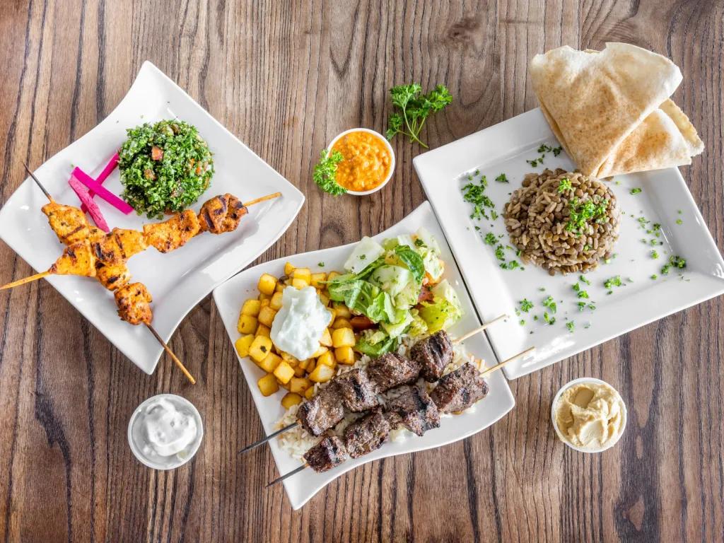 EVOO: Fresh Mediterranean Kitchen Three square plates on a wooden table, one with chicken kebabs with vegetables, another with lamb kebab and potato chopped into small squares and a salad and last of all, a plate with brown rice mixed with beans. There are three small pots of sauces.