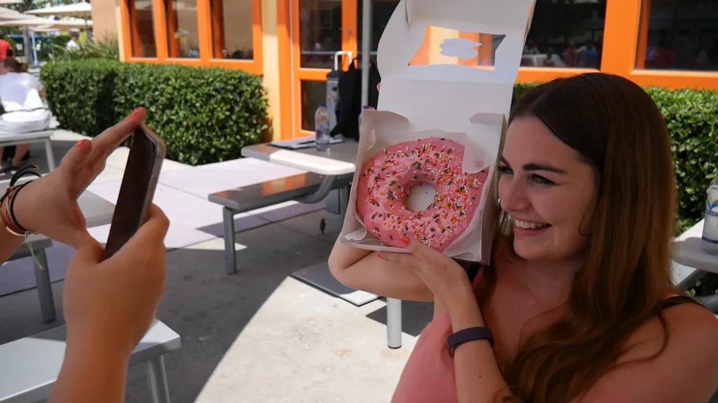 Lard Lad doughnut Someone taking a photo of a woman smiling holding a giant doughnut with pink icing and sprinkles on it. The doughnut is in a white paper box which she is holding up to the right of her head.
