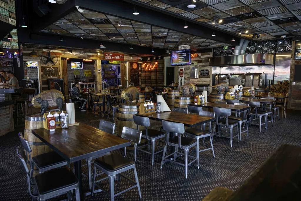 Brother Jimmy’s BBQ Darkly lit restaurant with black metal chairs surrounding dark wooden square tables with southern American country theming. There are barrels and neon signs on the wall with a tv playing sports events.