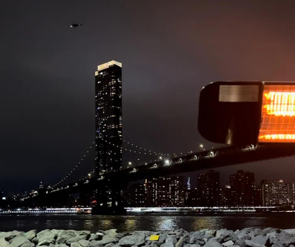 Cecconi's Dumbo To the right there is a heater and to the left there is an incredible view from a restaurant with skyscrapers and the Brooklyn Bridge lighting the night sky. There is also the river below and above a helicopter is flying.