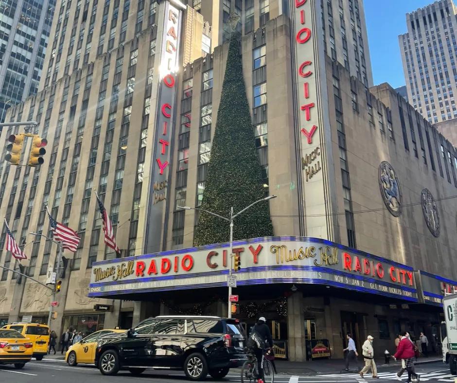 Radio City Music Hall Cars driving along the road below building with silver, red and blue sign saying "Radio City Music Hall'. There is a giant half christmas tree on top of the sign leaning against the wall. On the right there are American flags waving in the wind.