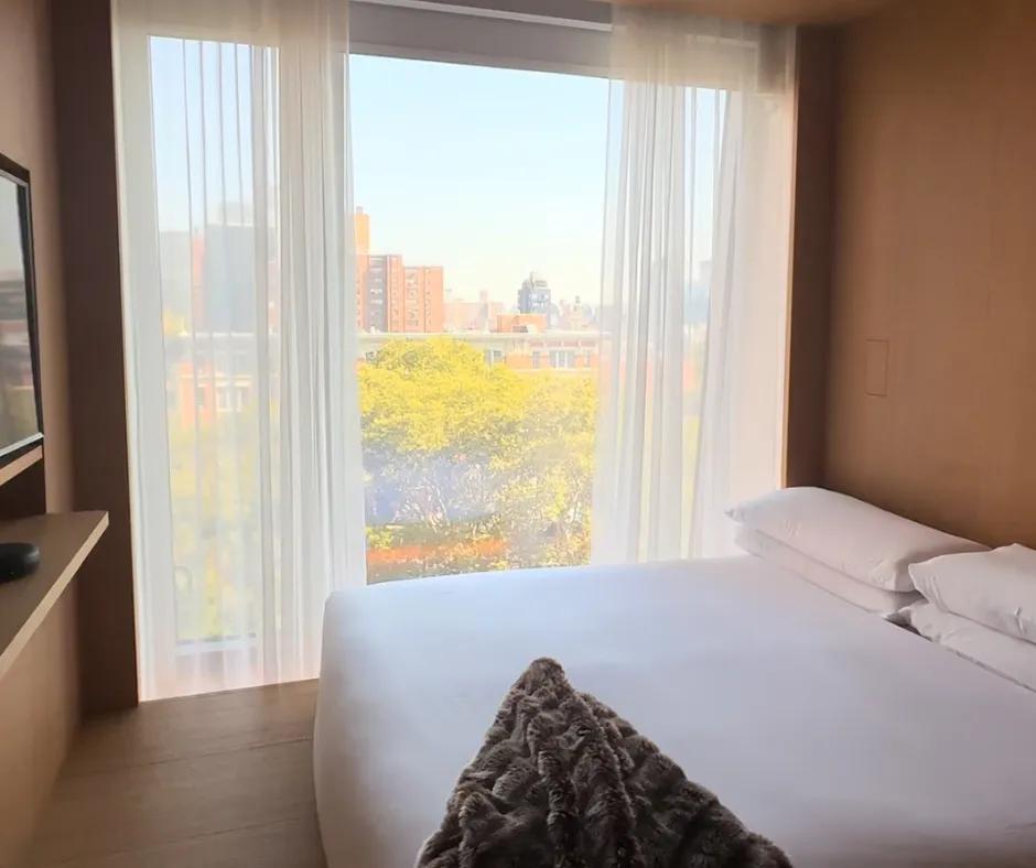PUBLIC Hotel, New York City Bed on wooden floor with white sheets next to a large window overlooking New York City. There is a television on the wall in front of the bed and white curtains slightly drawn closed.