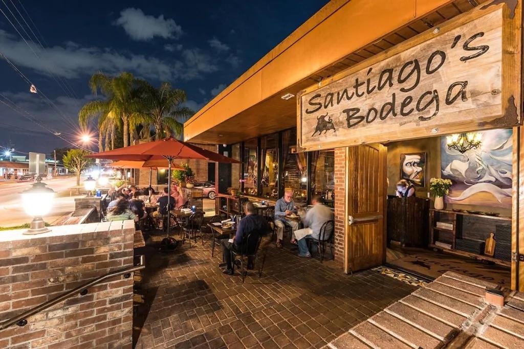 Santiago's Bodega Outside of a restroom with grey brick terrace and people sat beneath red umbrellas. There is a wooden sign which says Santiago's Bodega in black.