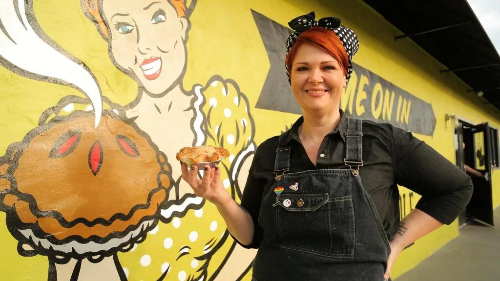Se7en Bites Woman with red hair stood holding pie in front of building with yellow and black art piece painted on side of woman holding a steaming hot pie.
