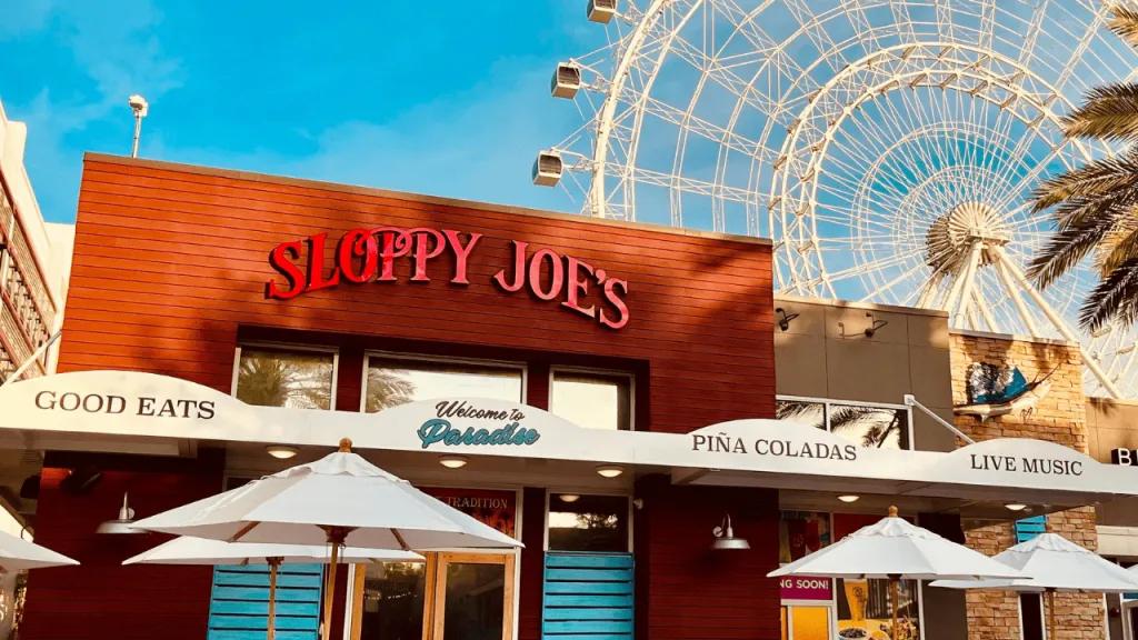 The Orlando Eye Box shaped restaurant coloured red and beige with a lighter red sign reading "Sloppy Joe's" with the white Orlando Eye in the background.