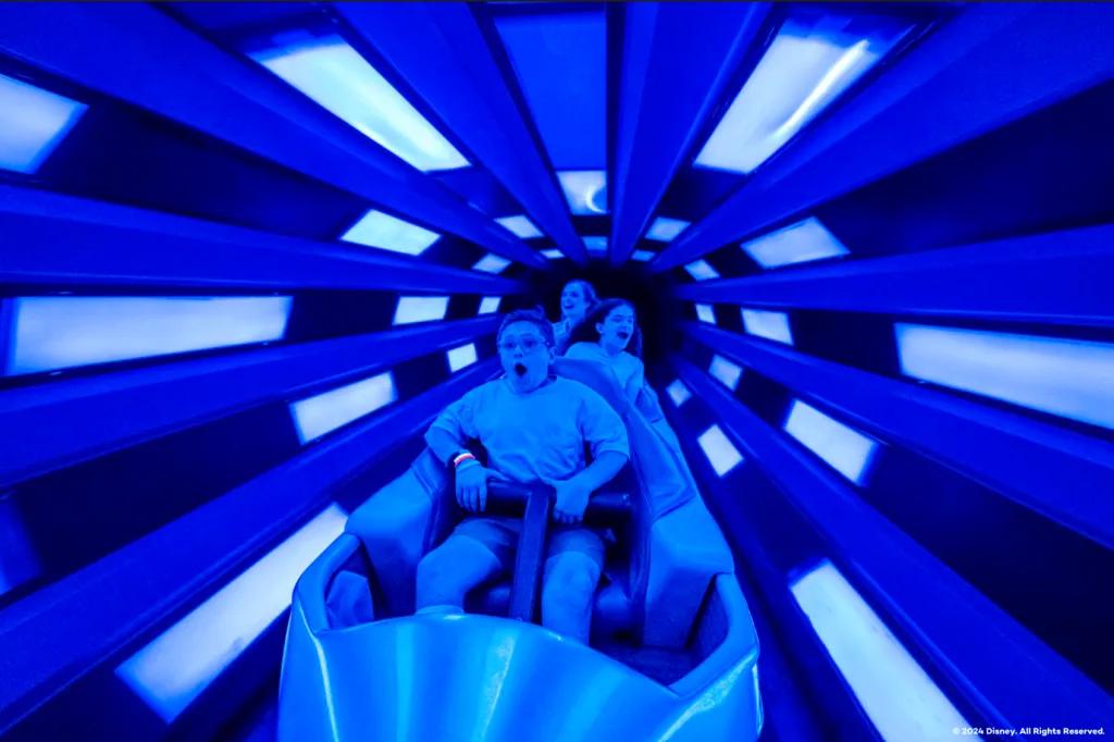 Space Mountain Three people sat one in front of the other on a rollercoaster vehicle in a tunnel lit blue