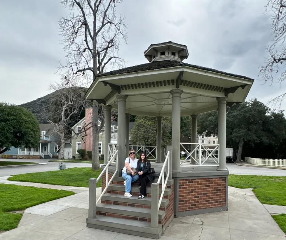 Stars Hollow gazebo Gazebo in the centre of a town with while pillars, a brick staircase and grey outlines. In the background there are tall trees without leaves on them, a mint green and white house and a blue house with white fences.