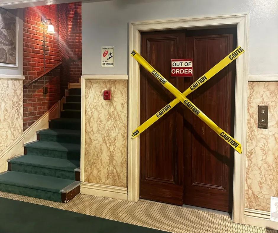 The Big Bang Theory apartment set Apartment staircase with brick walls, dark blue staircase and a beige and white wallpaper. There is a lift with a dark wooden door, an 'OUT OF ORDER' sign on the doors and two strips of caution tape in an X shape on the door.