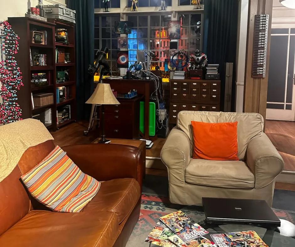 Sheldon's apartment set Apartment designed set, with dark brown sofa with patterned blanket on top and stripey cushion on. Another smaller sofa which is grey with an orange cushion. In the background there are DNA structure statues, comic books spread out on coffee table and robots on dark wooden drawers. To the right there is a white door with glass.