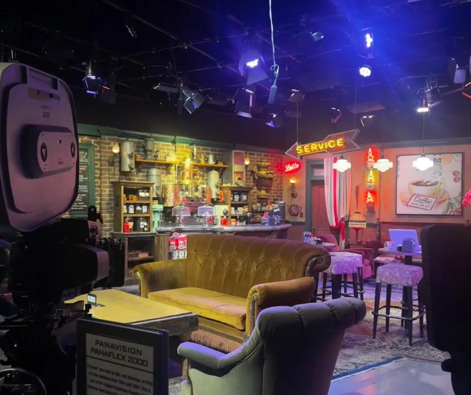 Central Perk set Set of the Friends coffee shop, to the left there is a film camera with the iconic coffee table and brown sofa behind. There is also the coffee counter with cake holders and signs saying 'service' hanging up. Behind there are other tables and some stools.
