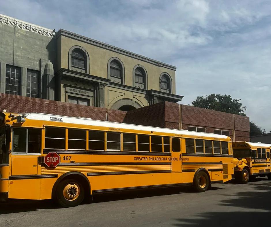 School buses on set Two large yellow school buses with a stop sign attached to one and writing along in black saying "GREATER PHILADELPHIA SCHOOL DISTRICT". In the background is a brick building and behind that is a couple more buildings.