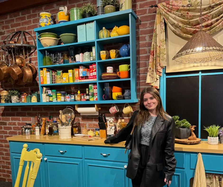 Friends kitchen set Woman in sparkly blue top and black blazer stood with hand on wooden kitchen counter that has light blue draws. Above there is cluttered utensils, condiments, pans hanging up and shelves with cutlery on.