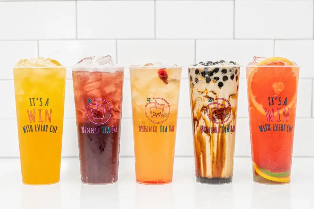 The Winnie Tea Bar Five large clear cups of different drinks, from left to right, there is a yellow drink with ice saying "It's a win with every cup", a dark red drink with ice with the logo on it, an orange drink with ice, a brown milk tea with small black tapioca pearls in and lastly a pink drink with slice of orange in.