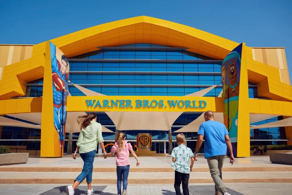 Warner Bros. World Family of four walking towards a large yellow building with two images either side of the entrance, one of superman comics and one of scooby doo. There is a blue sign saying "Warner Bros. World" and below there is a copper WB sign.